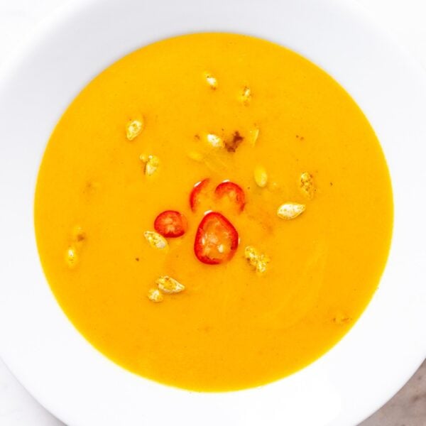 White bowl filled with curried butternut squash soup with toasted seeds and sliced red chiles on top all on white marble surface.