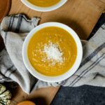 Top down view of bowl of curried butternut squash sitting on wood board with towel underneath with copper pot of extra soup and squash all on gray surface