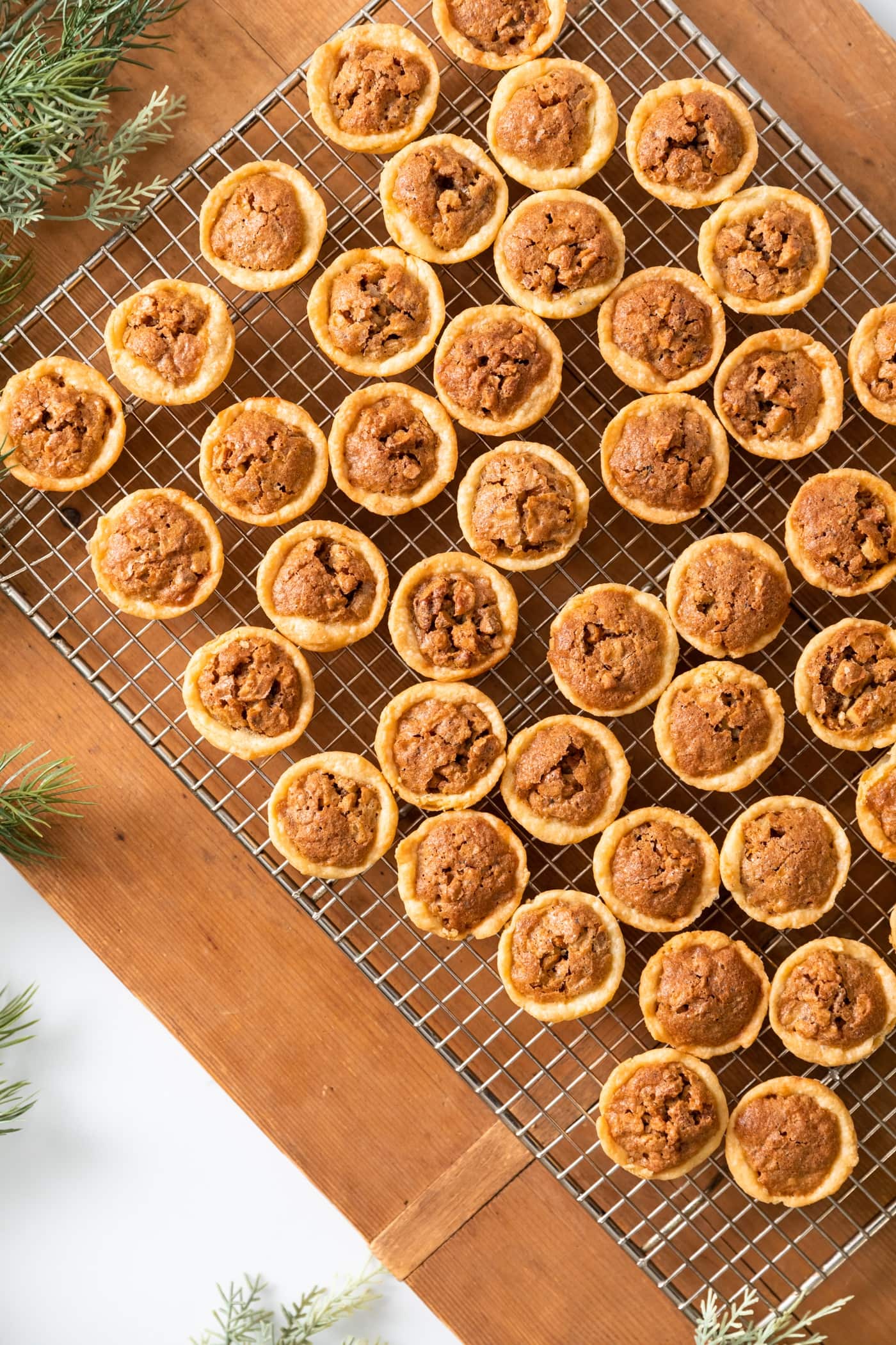 Top down view of many small pecan tassies sitting on cooling rack with wood board underneath with greens nearby