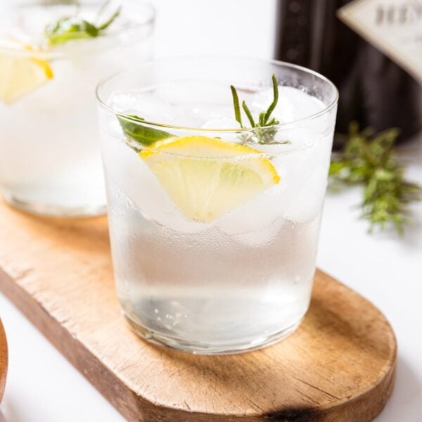 Two glasses filled with clear gin fizz liquid with lemon wedge and herbs sitting in glass sitting on wood board with small bowl filled with nuts.