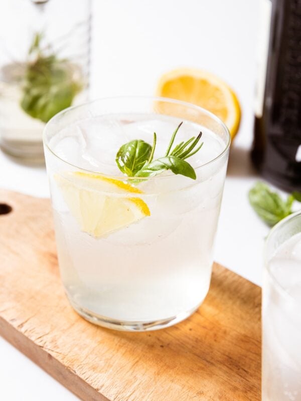 Glass filled with clear gin fizz with lemon wedge and herbs sitting on wood board with lemon in background.