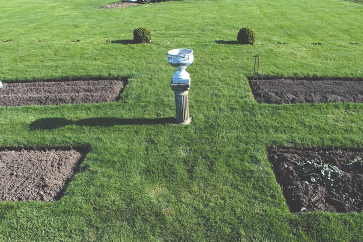 Large grass area with new garden beds dug out with a decorative urn on a pedestal in the center.