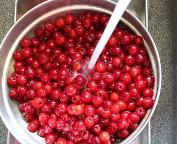 Large strainer filled with lots of pitted sour cherries with a stream of water running over the top to clean them.