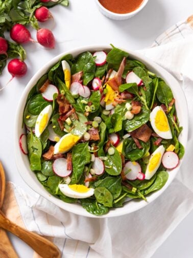 Top down view of spinach pieces in white bowl topped with bacon, eggs, and radish all on white countertop with extra radish