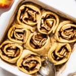 Top down view of white pan filled with apple dumpling rolls filled with cinnamon with one piece missing all on white countertop with apples