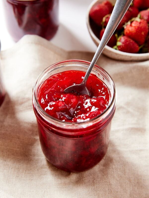 Jam jar filled with red colored strawberry jam sitting on towel with more strawberries behind.