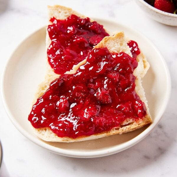 Strawberry jam recipe slathered on two slices of bread sitting on white plate with pieces of fruit visible in jam.