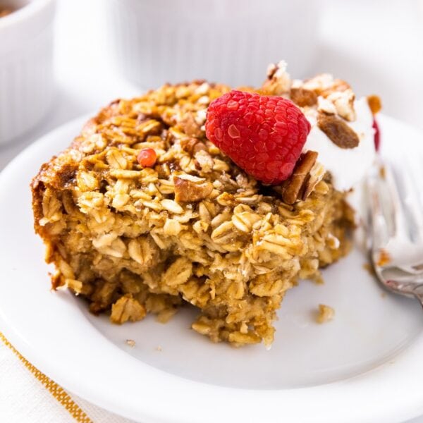 Square slice of baked oatmeal sitting on white plate with raspberry on top and fork beside with white napkin underneath