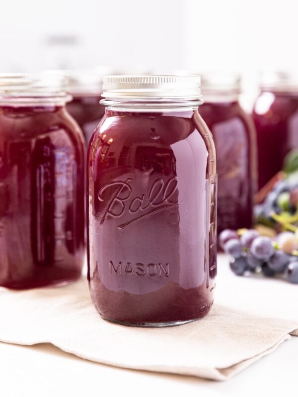 Glass jar filled with dark purple grape juice concentrate sitting on white towel with grapes in background behind as well as other glass jars