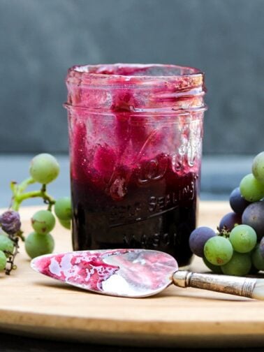Jar of grape butter sitting on cutting board with knife in front spread with grape butter and grapes to the side all on a piece of gray slate