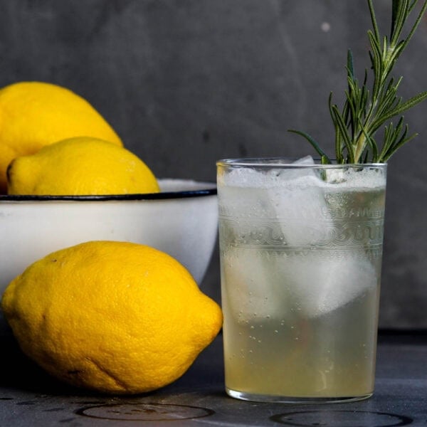 Glass of Venetian lemonade with ice and a sprig of rosemary sitting on blue slate surface beside of bowl of lemons