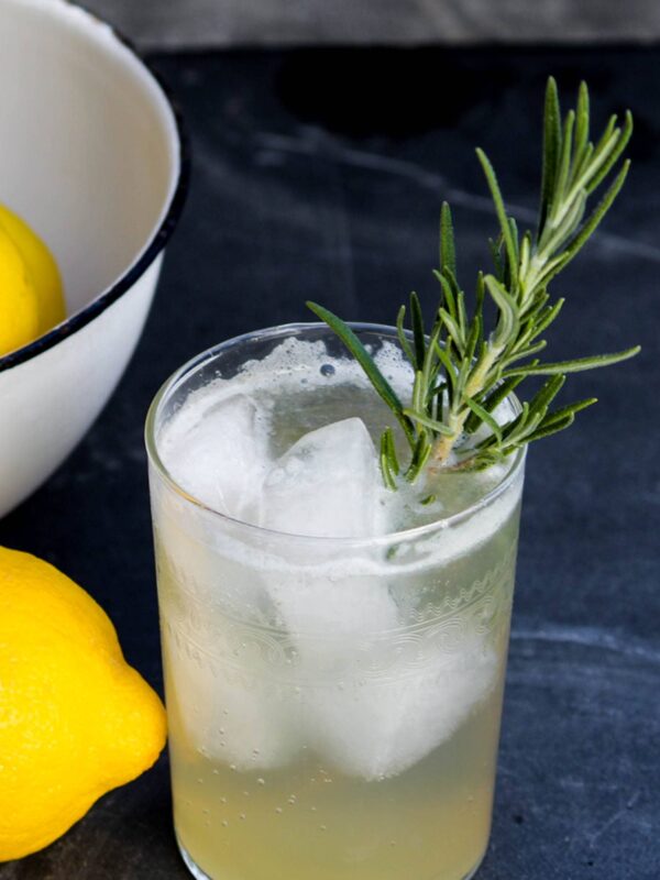 Glass of Venetian lemonade with ice and a sprig of rosemary sitting on blue slate surface beside of bowl of lemons