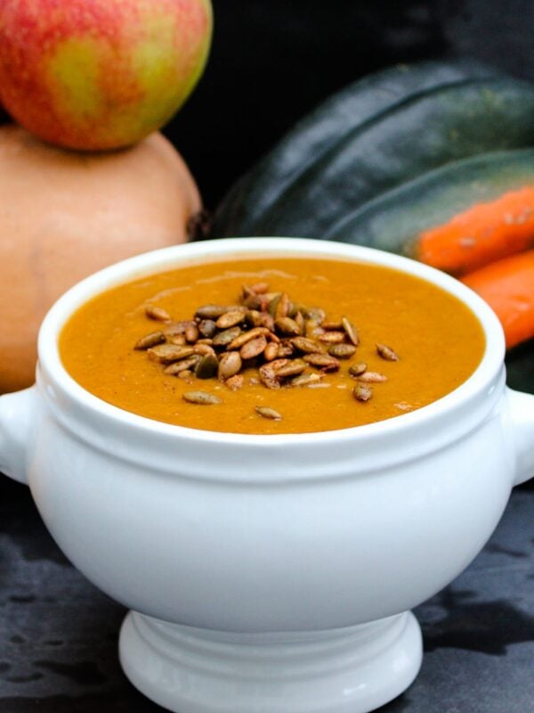 Bowl of orange colored harvest squash soup in white bowl with roasted pepitas on top with apple and various pumpkins and squash against a dark gray background