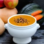 Bowl of orange colored harvest squash soup in white bowl with roasted pepitas on top with apple and various pumpkins and squash against a dark gray background