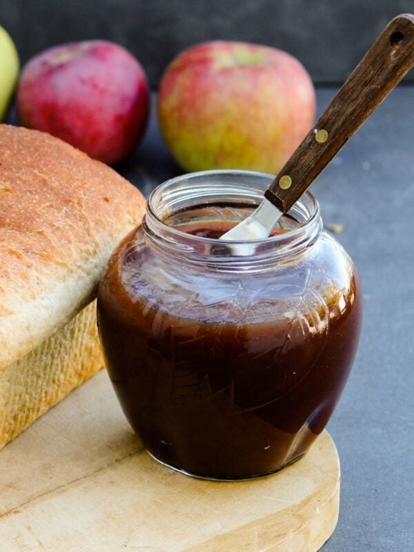 Jar of slow cooker apple butter with loaf of bread beside it and knife in front of a gray slate surface with extra apples in background