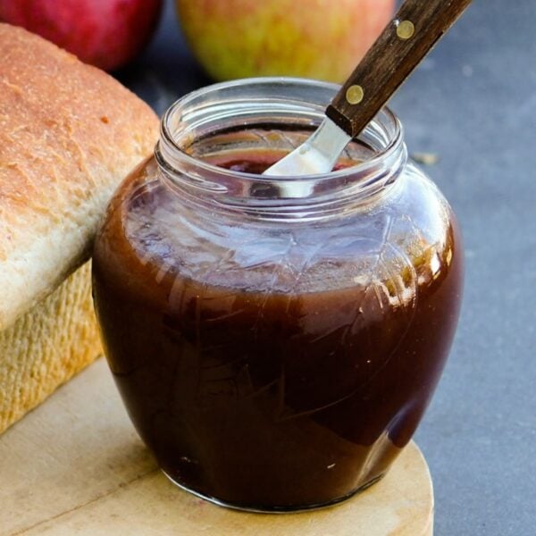 Jar of slow cooker apple butter with loaf of bread beside it and knife in front of a gray slate surface with extra apples in background