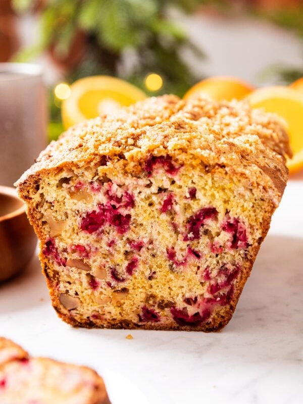 Cranberry orange bread with front sliced off showing interior texture of bread all sitting on white marble surface with Christmas lights in background.