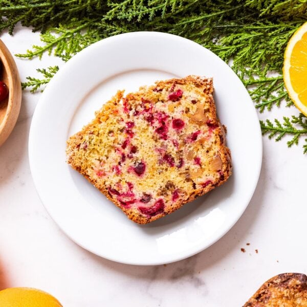White plate with slice of cranberry orange bread with cranberries and sliced oranges all around as well as green garland.