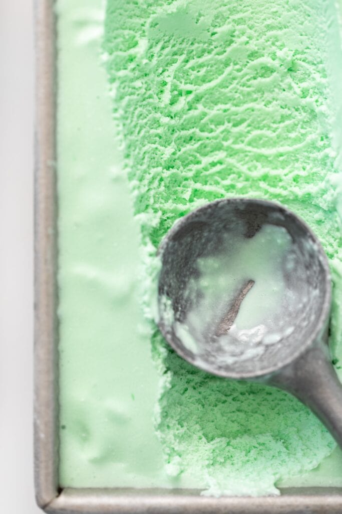 Top down view of ice cream scooper sitting on top of green colored ice cream in gray pan all on white surface
