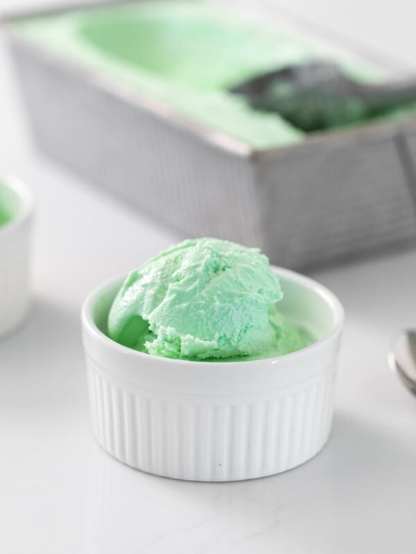 Green colored ice cream sitting in small white bowl with gray pan holding additional ice cream in background all on white surface