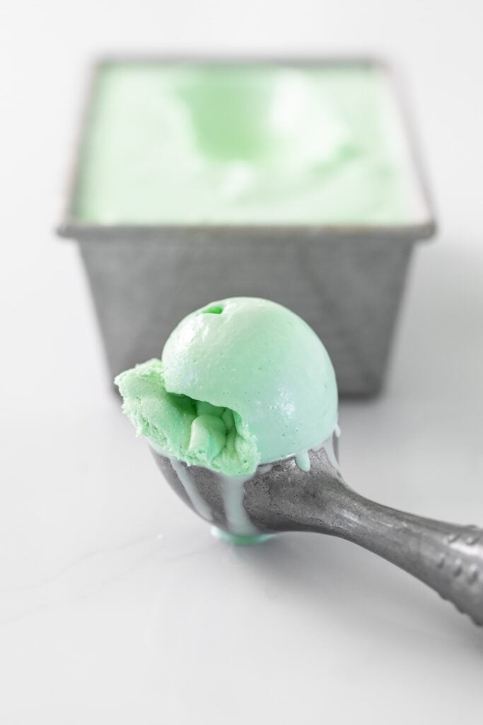 Gray ice cream scooper sitting on white countertop with green scoop of ice cream with gray pan in background