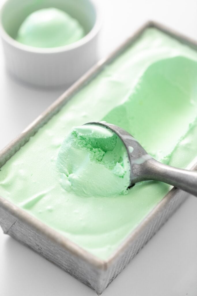 Ice cream scooper sitting in green colored ice cream in gray pan with small white bowl in background all on white surface