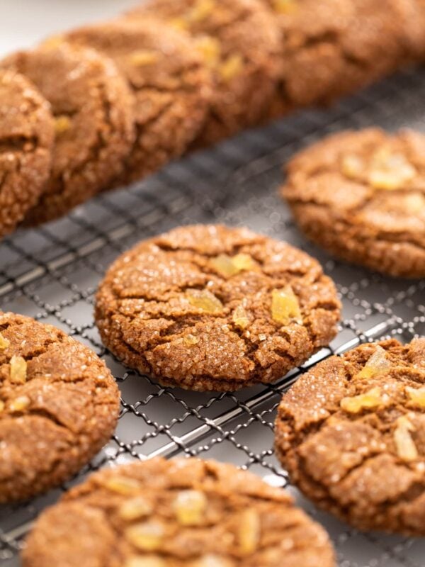 Brown colored molasses crinkle cookie sitting on cooling rack with other cookies stacked around all on white surface