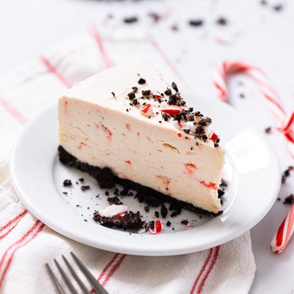 White plate sitting on white countertop with slice of peppermint cheesecake with chocolate crumbles on top with pieces of crushed up peppermint