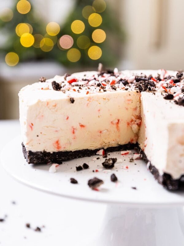 Side view of slightly pink peppermint cheesecake sitting on white cake plate with holiday twinkling lights in background