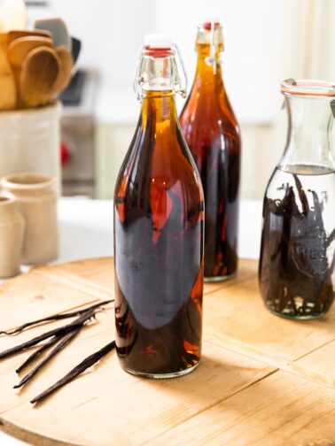 Large narrow bottle sitting on wood board filled with brown colored liquid with vanilla beans sitting beside with bottle with clear liquid behind