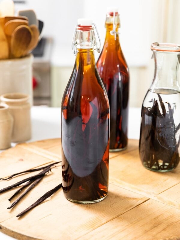 Large narrow bottle sitting on wood board filled with brown colored liquid with vanilla beans sitting beside with bottle with clear liquid behind