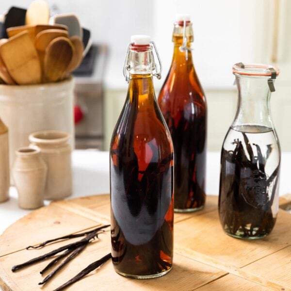Large bottle filled with brown liquid sitting on wood board with vanilla beans sitting beside with crock filled with wooden spoons in background