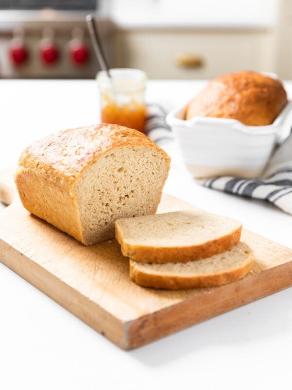 Loaf of sliced bread sitting on cutting board with jam container in background and unsliced loaf in white bread container all on white countertop