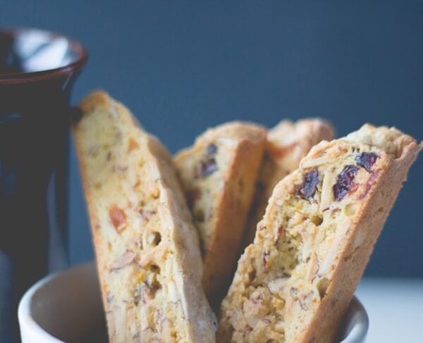 White bowl filled with slices of fruit and nut biscotti dotted with pieces of dried fruit with coffee cup in background