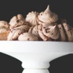 White cake plate filled with espresso chocolate meringue cookies with chocolate flecks all over in front of black background.