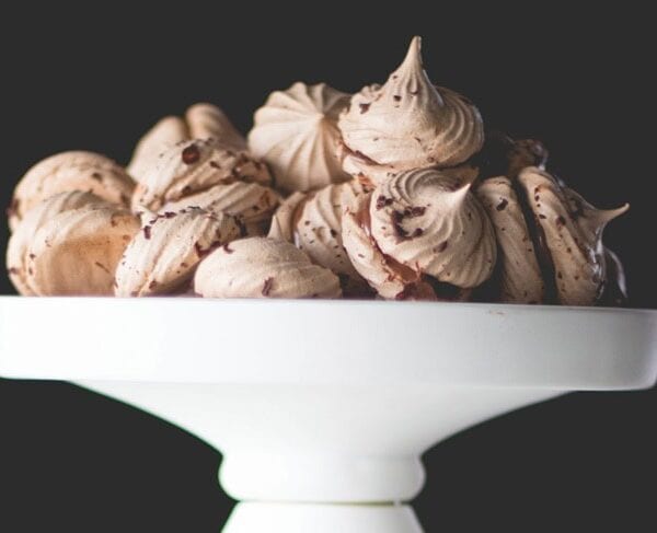 White cake plate filled with espresso chocolate meringue cookies with chocolate flecks all over in front of black background.