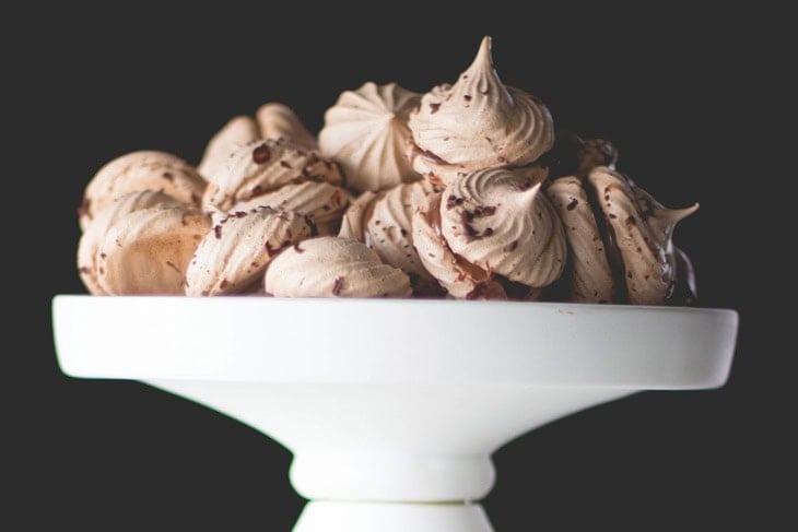 White cake plate filled with espresso chocolate meringue cookies with chocolate flecks all over in front of black background.