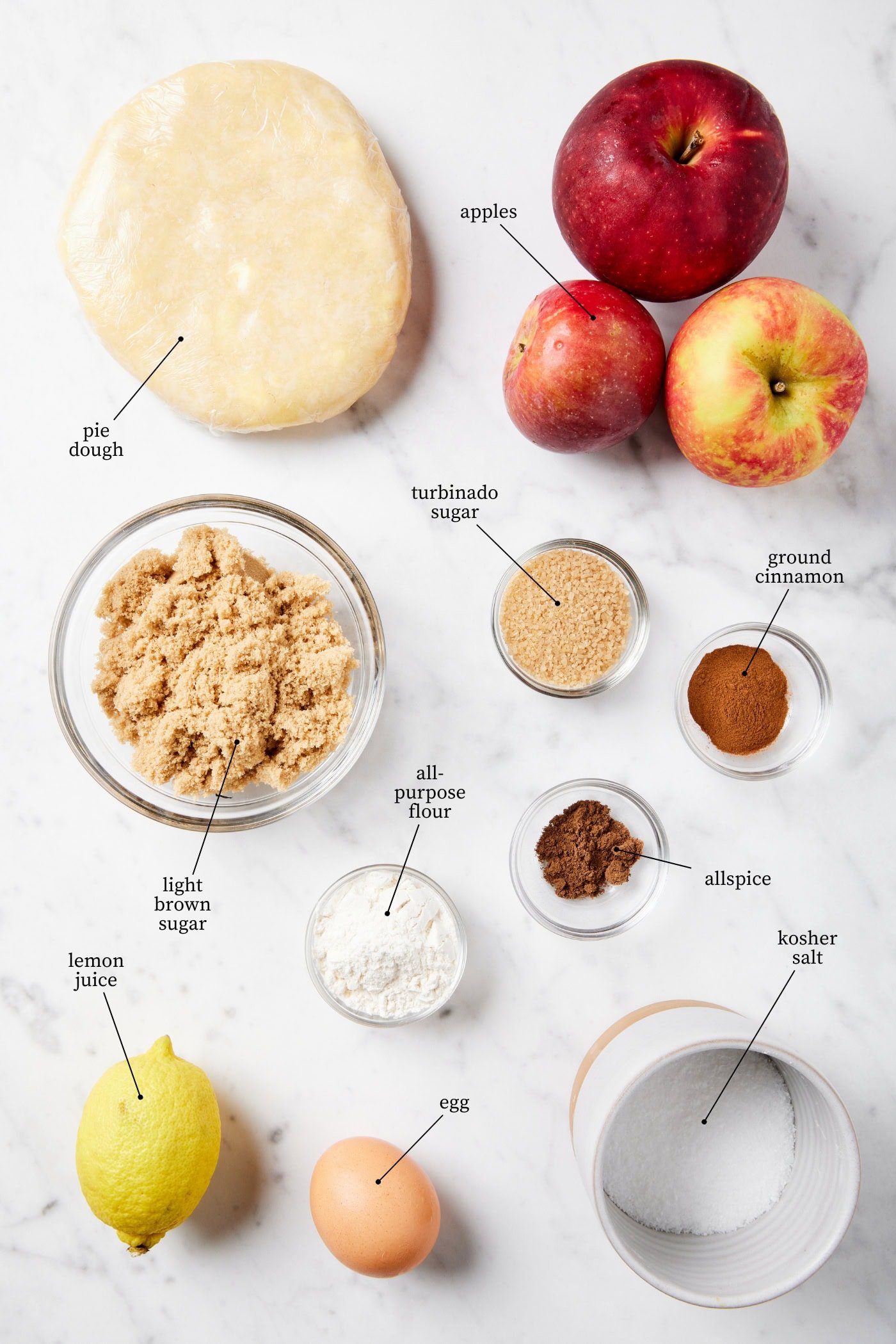 White marble surface filled with ingredients to make a rustic apple galette recipe including dough, apples, sugar, and more.