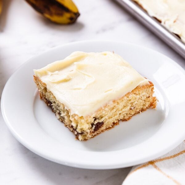 White plate with serving of banana bar with cream cheese frosting on top with rest of banana bars in background in metal baking pan.