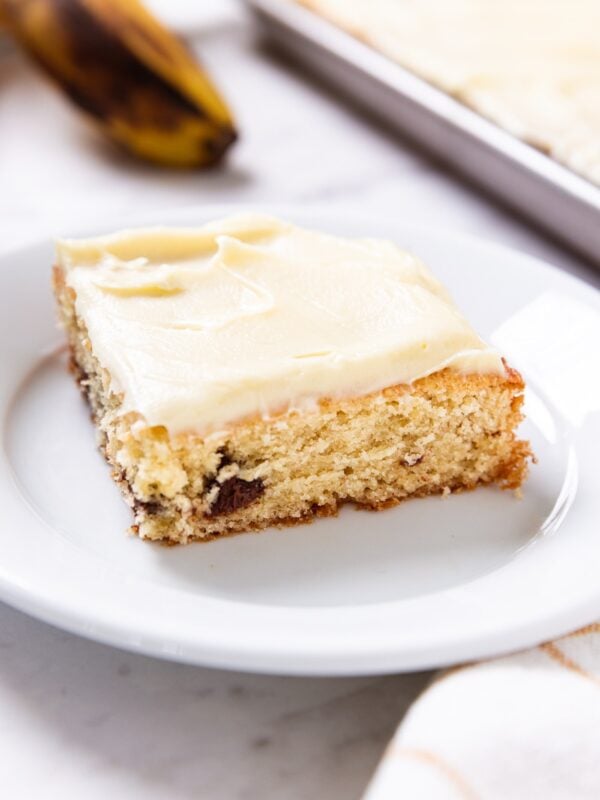 White plate sitting on marble surface with square of chocolate chip banana bar sitting on plate with banana in background along with rest of banana bars.