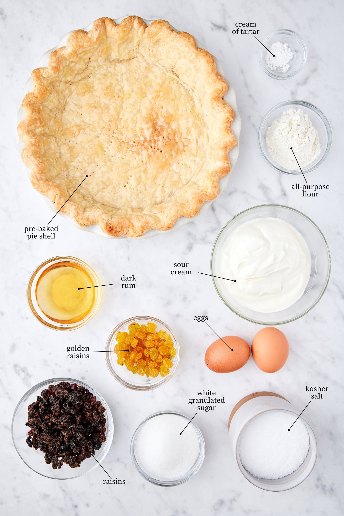 White marble surface filled with ingredients needed for a sour cream raisin pie including pie crust, raisins, sugar, flour, rum, and more.