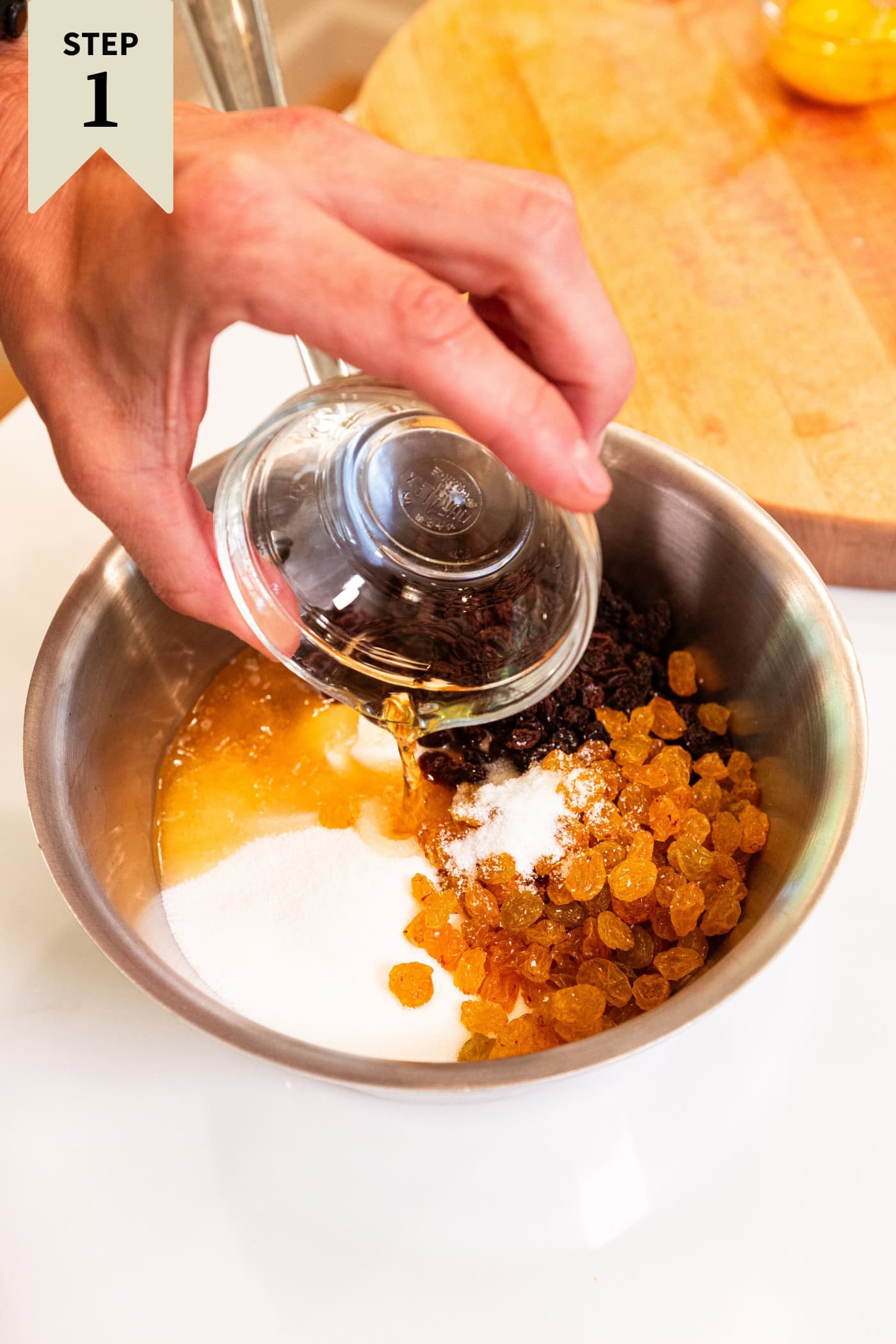 Hand pouring rum ingredients into a metal saucepan filled with raisins, sugar, and sour cream.
