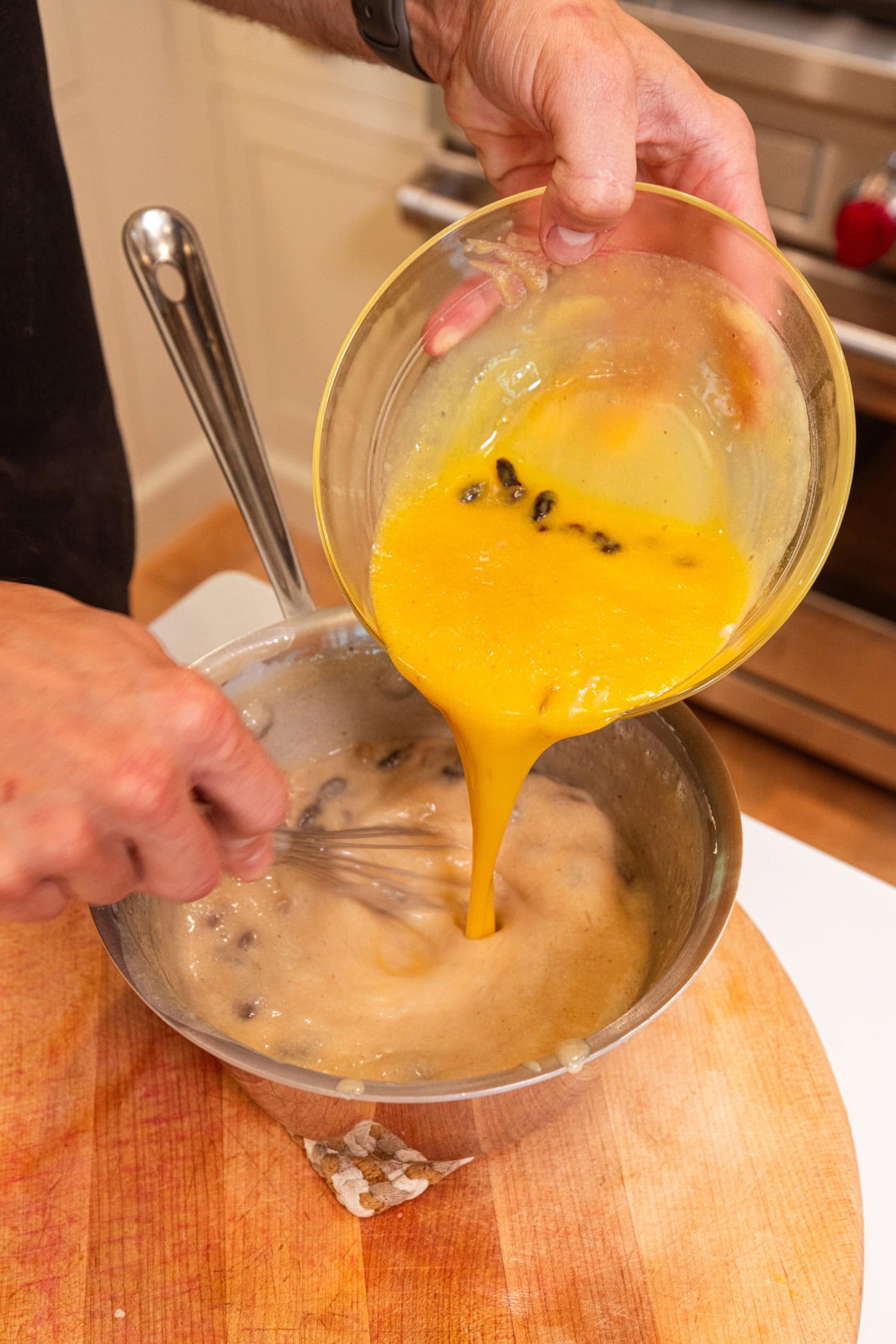 Glass bowl with some tempered egg yolks being poured back into a saucepan with rest of mixture.