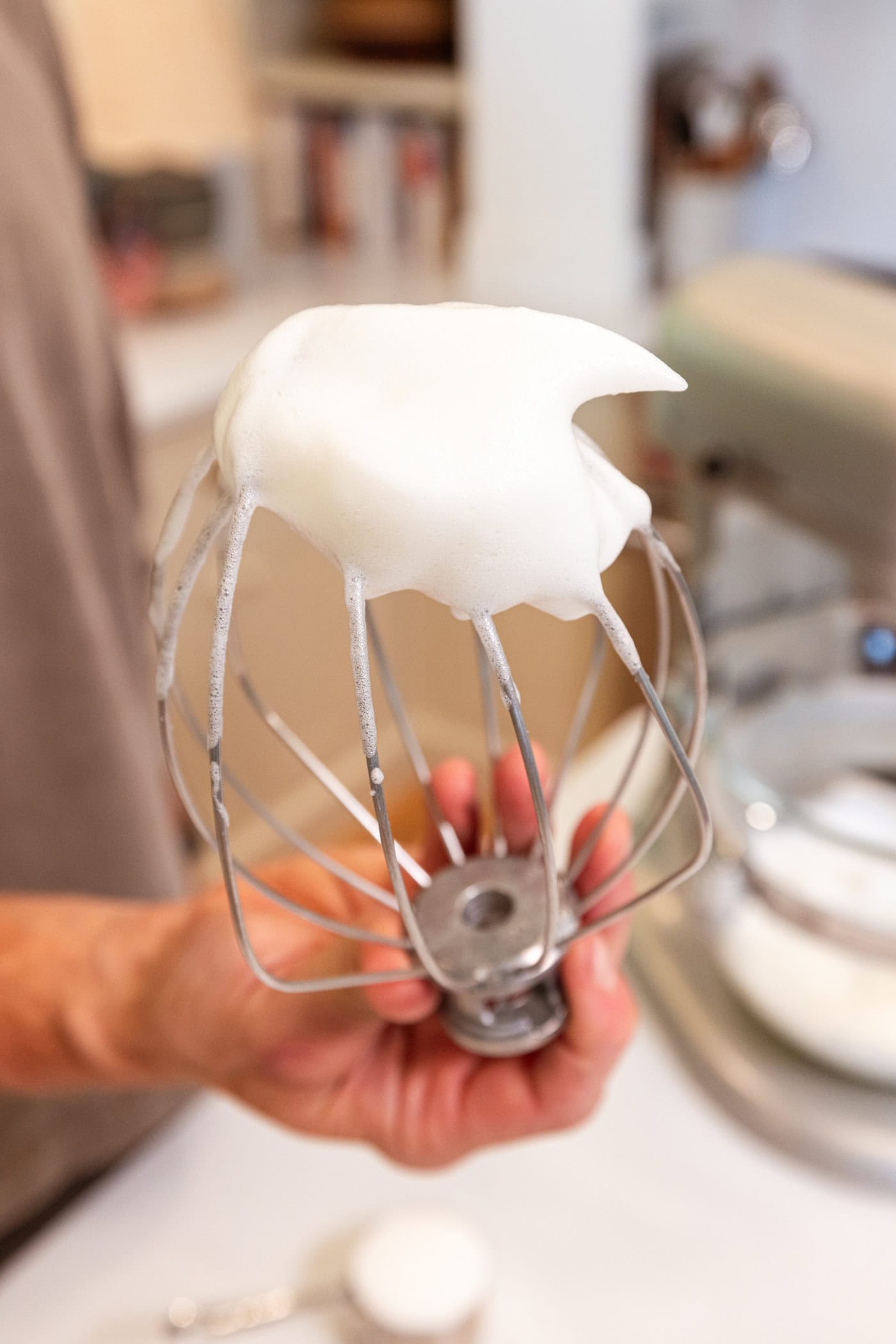Hand holding whisk attachment to stand mixer to show the fluffy texture of whipped egg whites.