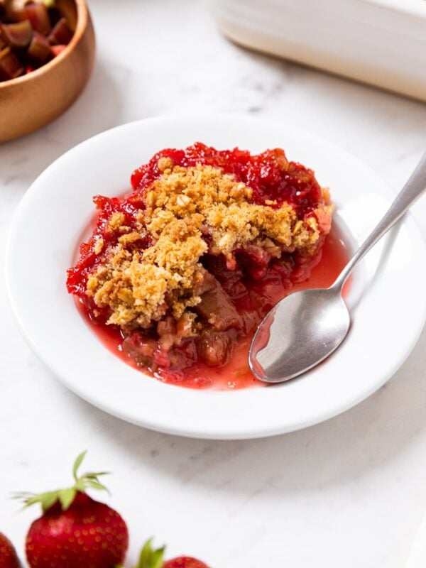 White plate sitting on white marble surface with serving of strawberry rhubarb crumble topped with streusel topping with strawberries and rhubarb and rest of crisp around.