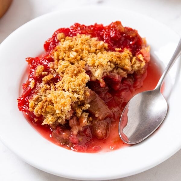 Small white plate filled with serving of strawberry rhubarb crumble with spoon resting to the side after taking a bite with baking dish filled with rest of crumble in the background.