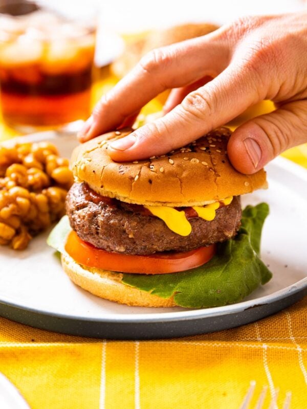 Hand pushing down on top bun of grilled burgers sitting on white plate with baked beans to the side with mustard dripping down the side of the hamburger