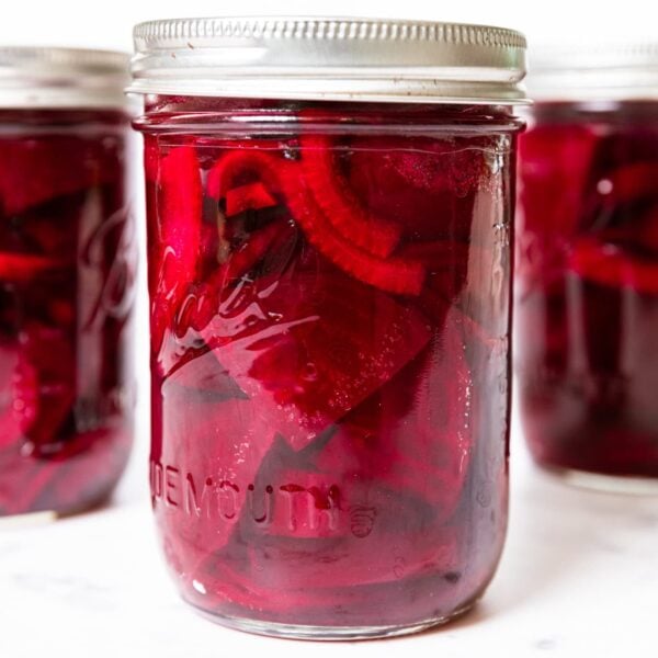 Cans of pickled beets with onions in red liquid sitting on marble surface with lids and rings.