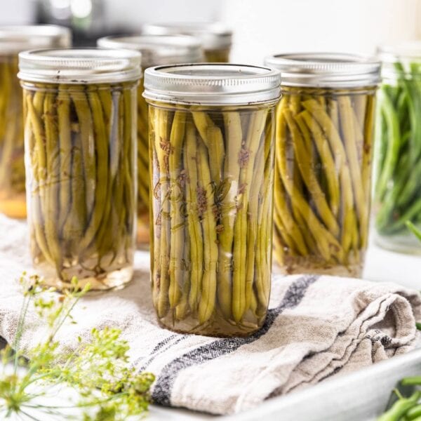 Glass canning jars filled with long green beans with extra glass jars in background and some fresh green beans in foreground