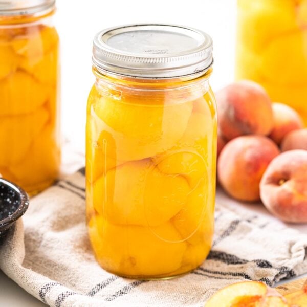 Large glass jar filled with slices of peaches sitting on white countertop with extra glass jars around as well as unsliced peaches